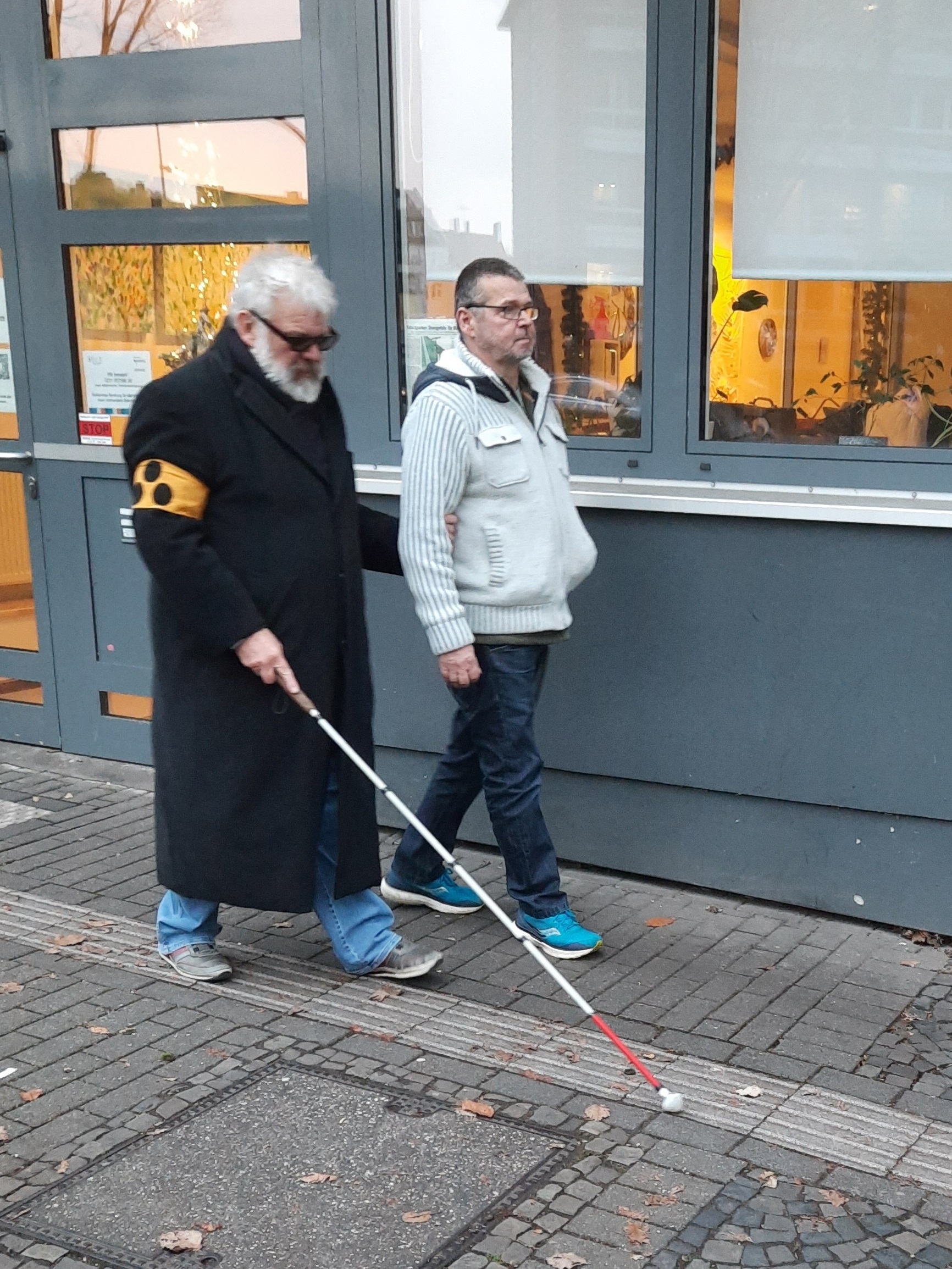 Ein sehender Helfer führt einen blinden Mann auf dem Gehweg